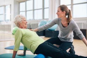Physical therapist working with a senior woman at rehab. Female trainer helping senior woman doing exercise on foam roller at gym.