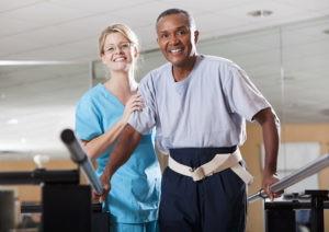 A female physical therapist wearing blue scrubs is working with a senior, African American male patient, doing gait training. The man is holding onto parallel bars for balance and is wearing a belt that the therapist can hold onto to help him do his walking exercises. They are looking at the camera, smiling.
