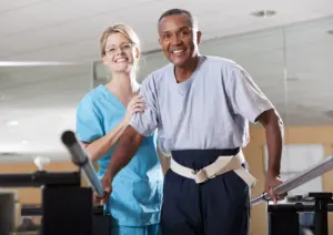 A female physical therapist wearing blue scrubs is working with a senior, African American male patient, doing gait training. The man is holding onto parallel bars for balance and is wearing a belt that the therapist can hold onto to help him do his walking exercises. They are looking at the camera, smiling.